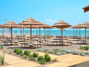 A beachside area with rows of sun loungers and thatched umbrellas, a wooden deck, and a view of the ocean.