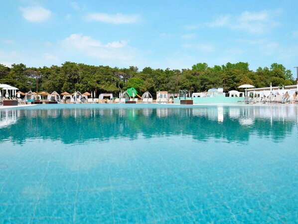Piscina azul cristalina com espreguiçadeiras e guarda-sóis, cercada por árvores e um edifício com fachada azul clara.