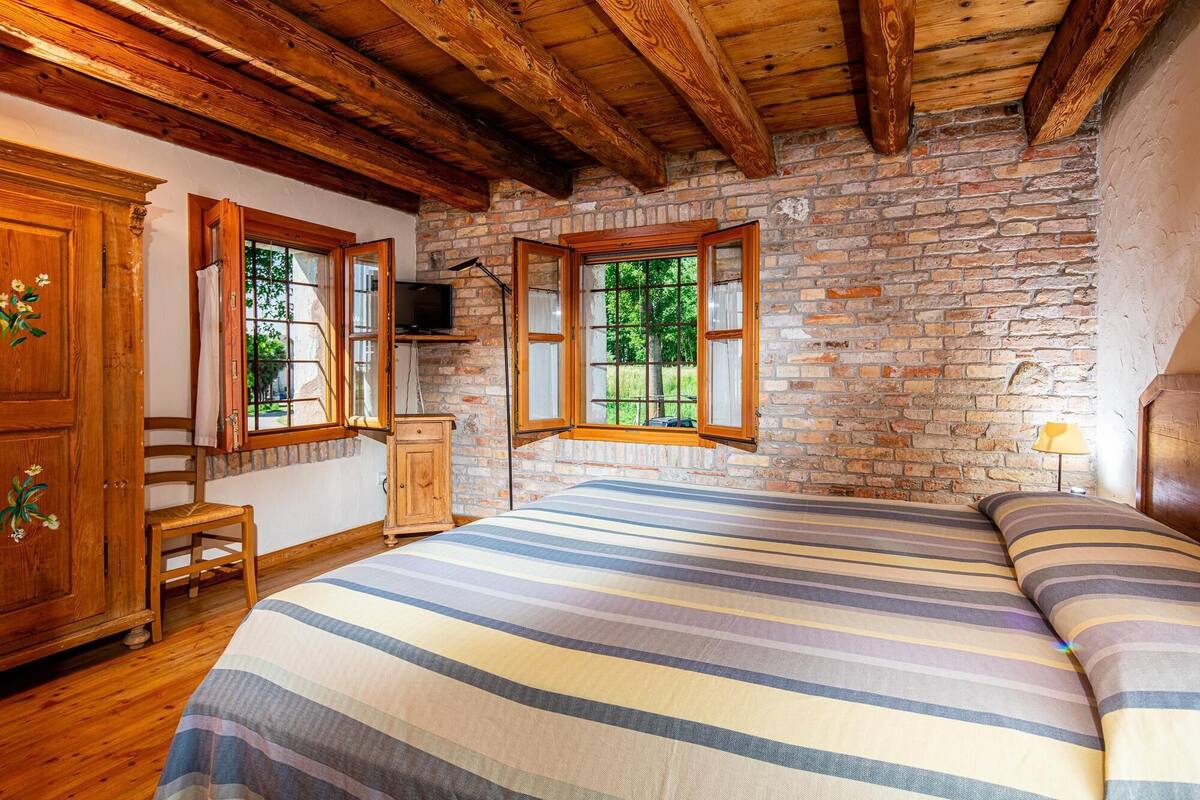 A rustic bedroom with exposed brick walls, wooden beams, a bed with striped bedding, a wooden cabinet, and a small table.
