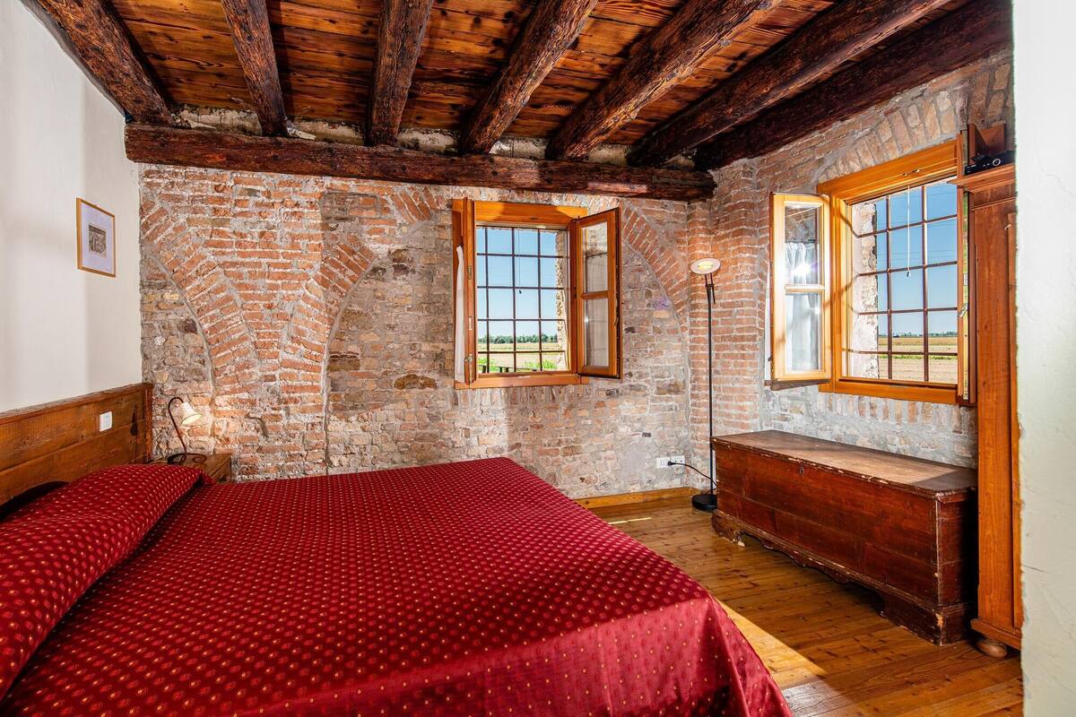 A rustic room with exposed brick walls, wooden beams, and a large bed with a red patterned bedspread.