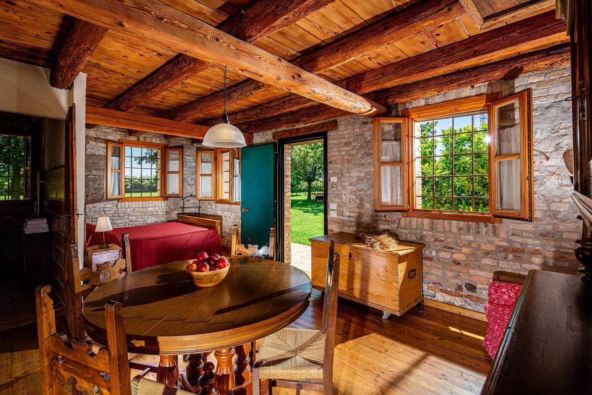 A rustic wooden interior with a stone wall, a round wooden table with chairs, and a view of greenery outside.