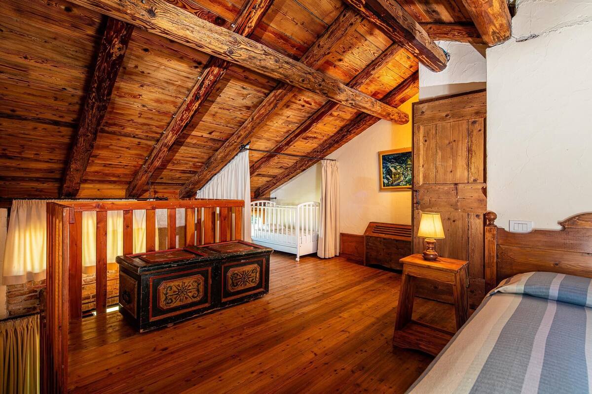 A rustic room with exposed wooden beams, a crib, a bedside table with a lamp, and a bed with a striped bedspread.