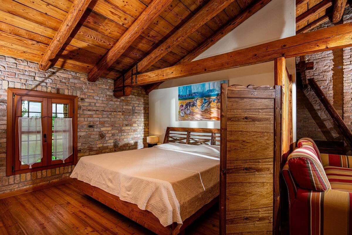 A rustic bedroom with exposed wooden beams, a brick wall, a bed with a white bedspread, a painting on the wall, and a striped chair.