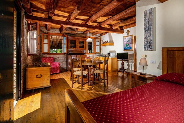 Ein rustikaler Raum mit freiliegenden Holzbalken, einem Holzesstisch mit Stühlen, einem rot gepolsterten Sessel, einer hölzernen Kommode, einem Bett mit roter Bettdecke, einem Fernseher und einem an der Wand befestigten Kruzifix.