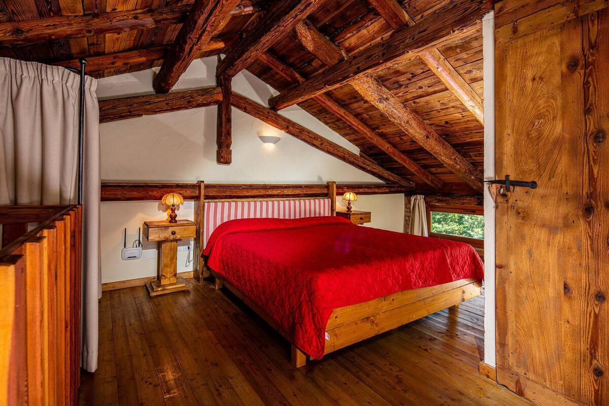 A wooden-framed bedroom with a red bedspread, two bedside lamps, and a window with a view of greenery.