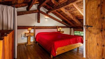Ein schlicht eingerichtetes Schlafzimmer mit Holzbettrahmen, roter Bettdecke, zwei Nachttischlampen und einem Fenster mit Blick ins Grüne.