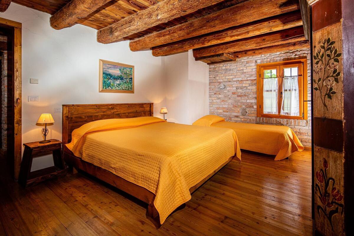 A room with two beds, wooden flooring, exposed wooden beams, a brick wall, a window with curtains, and a painting on the wall.