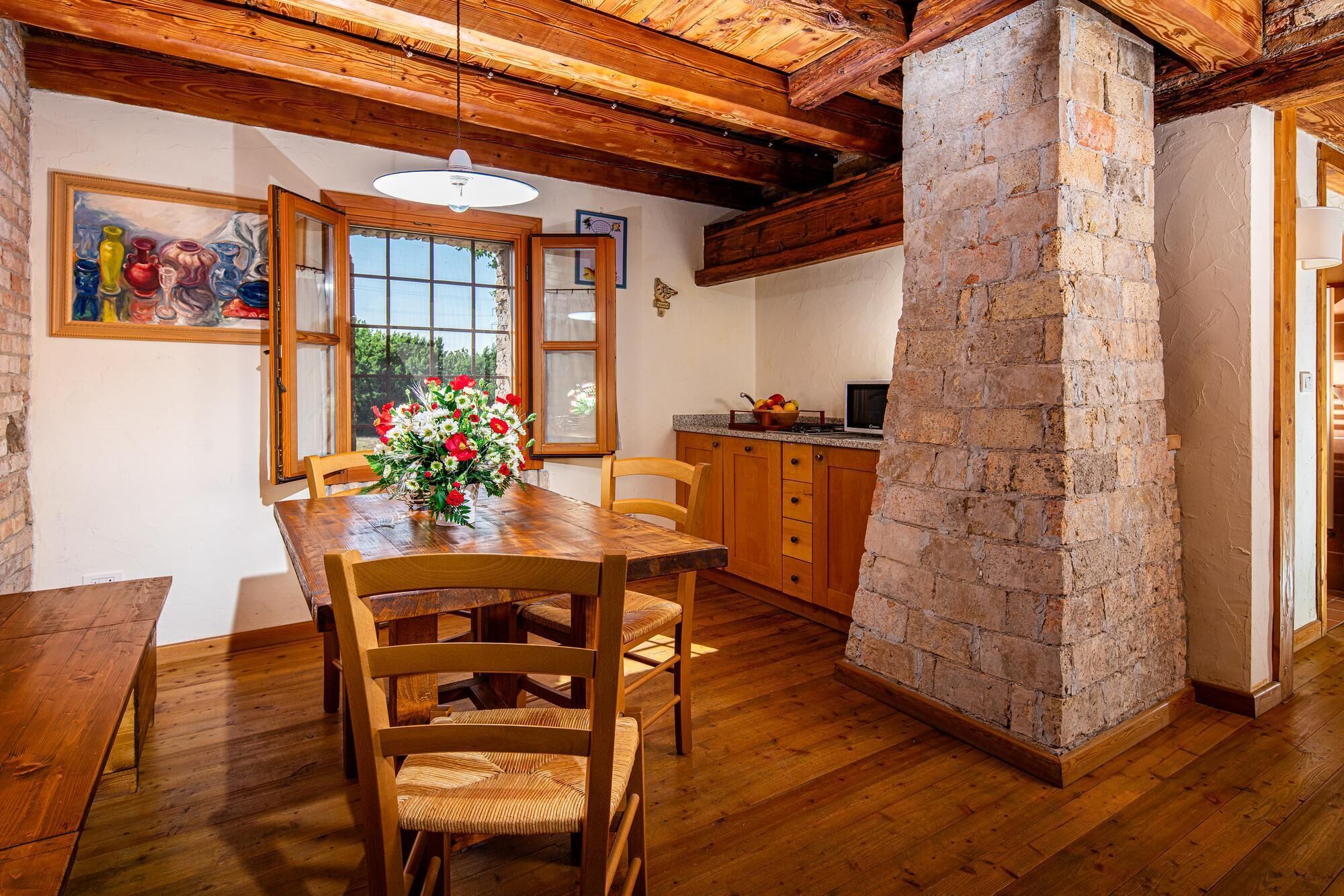 A dining area with a wooden table and chairs, a brick column, and a kitchen in the background.