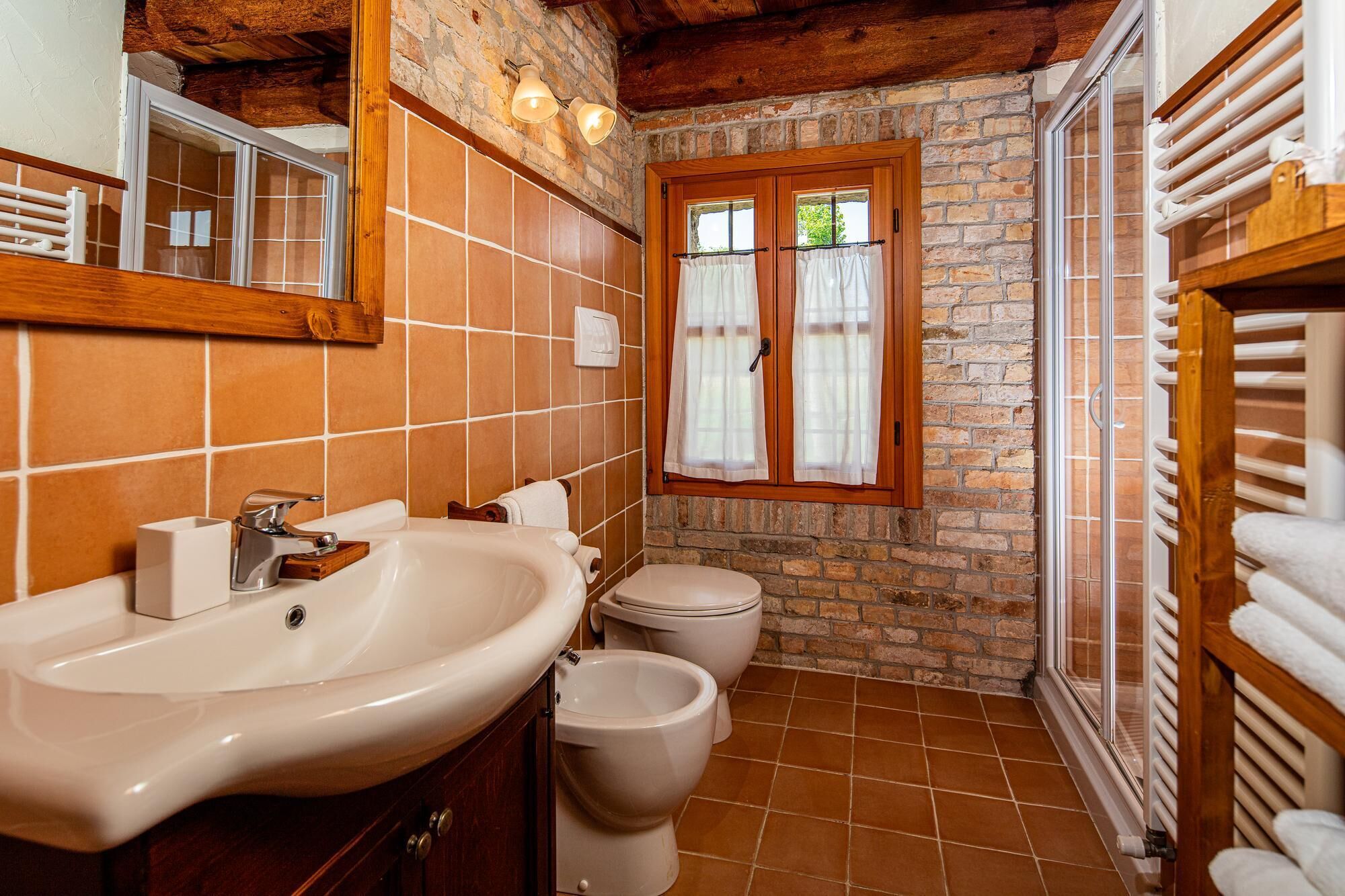 A bathroom with a stone wall, a wooden ceiling, a large sink, two toilets, and a window with curtains.
