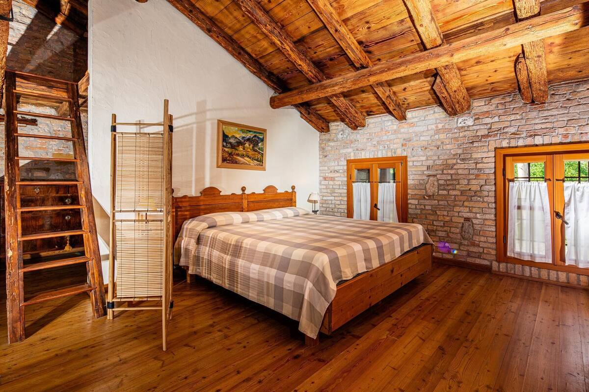 A bedroom with a wooden bed, a ladder, a painting, and a brick wall.