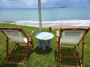 Two wooden chairs with mesh backrests and a small round table, set on a grassy area with a view of the ocean and distant islands.