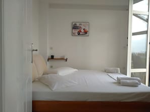 A bedroom with a bed, pillows, a small shelf with items, and a window. - Alekos Beach Houses-Roni (Kefalonia)