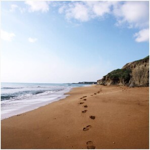 A sandy beach with footprints leading to the water, a cliff on the right, and a clear sky.