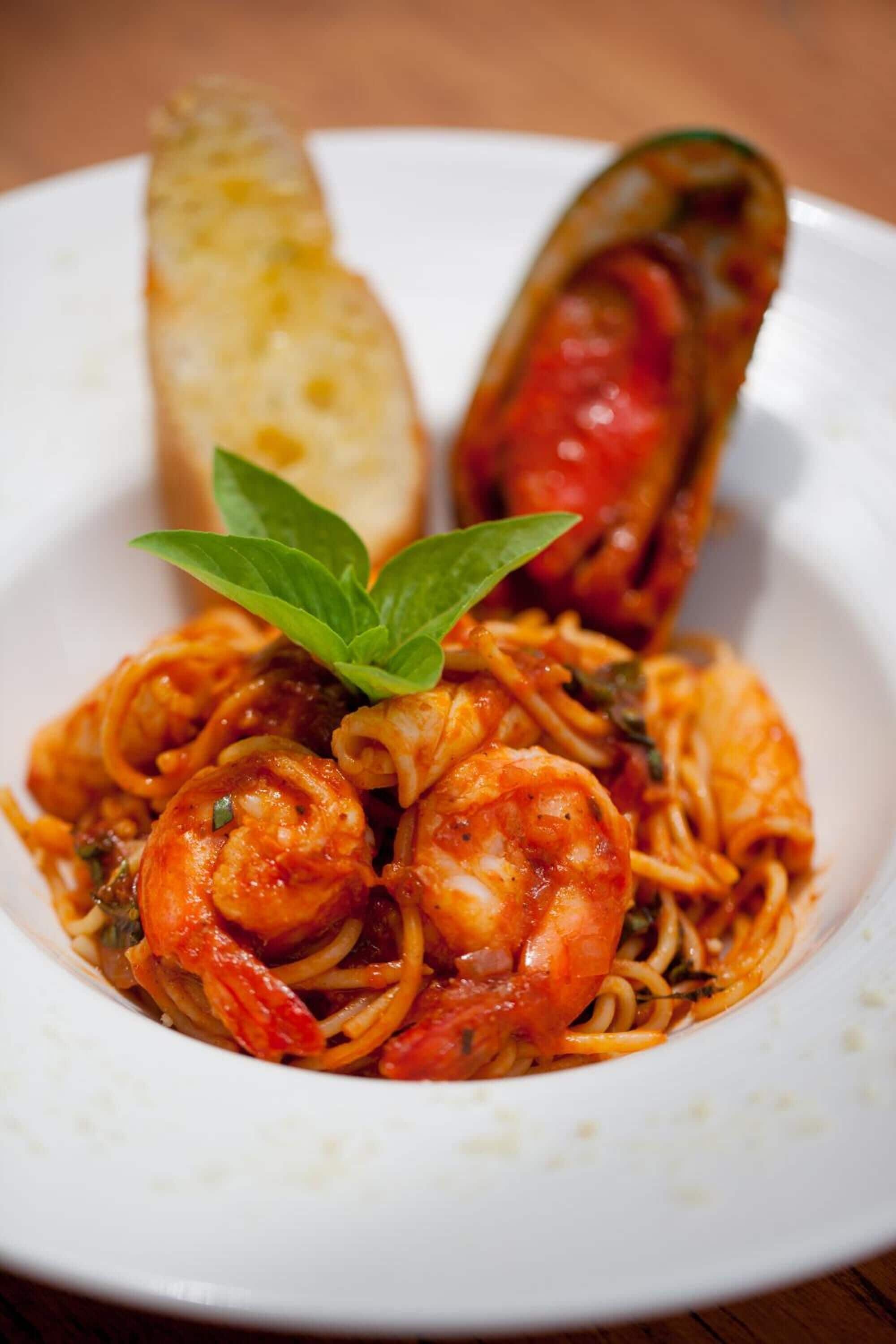 Un plato de espagueti con camarones y mejillones, adornado con una hoja de albahaca, servido con una rebanada de pan.