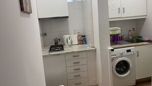 Una cocina compacta con armarios blancos, cocina a gas, hervidor, lavadora y un lavamanos pequeño.