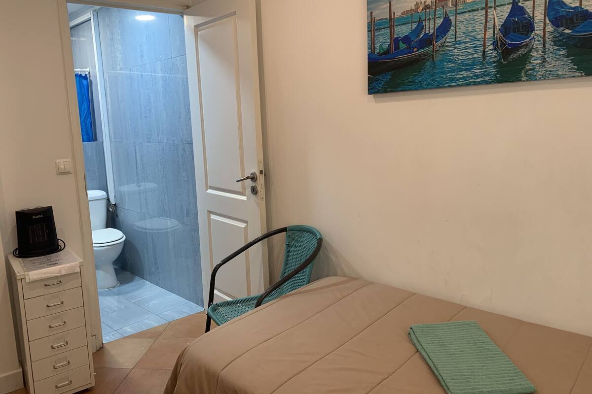 A room with a bed, a chair, a toilet, and a painting of boats on a wall.