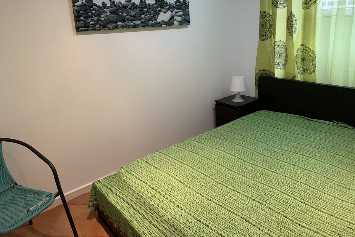 A bedroom with a bed, a chair, a nightstand with a lamp, a painting of a rocky beach, and a window with patterned curtains.