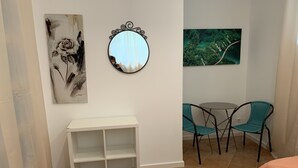Una habitación con una cama, un pequeño estante blanco, dos sillas azul verdosas, un espejo redondo y dos cuadros.