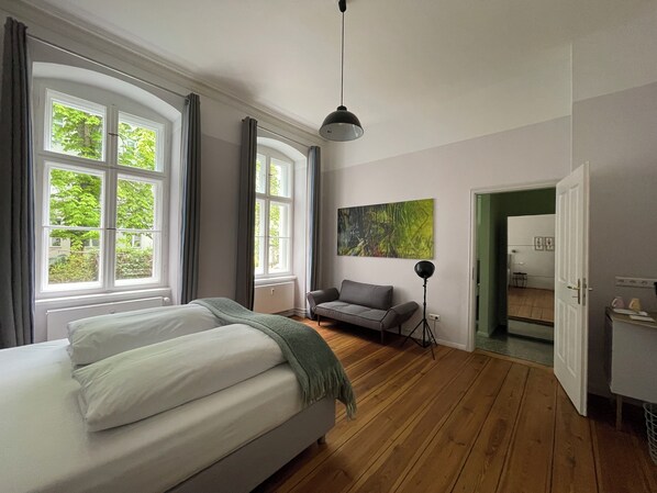 A bedroom with a bed, a gray sofa, a floor lamp, and a painting on the wall.