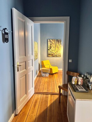 A room with a wooden floor, a yellow chair, a painting on the wall, and a door with a sign.