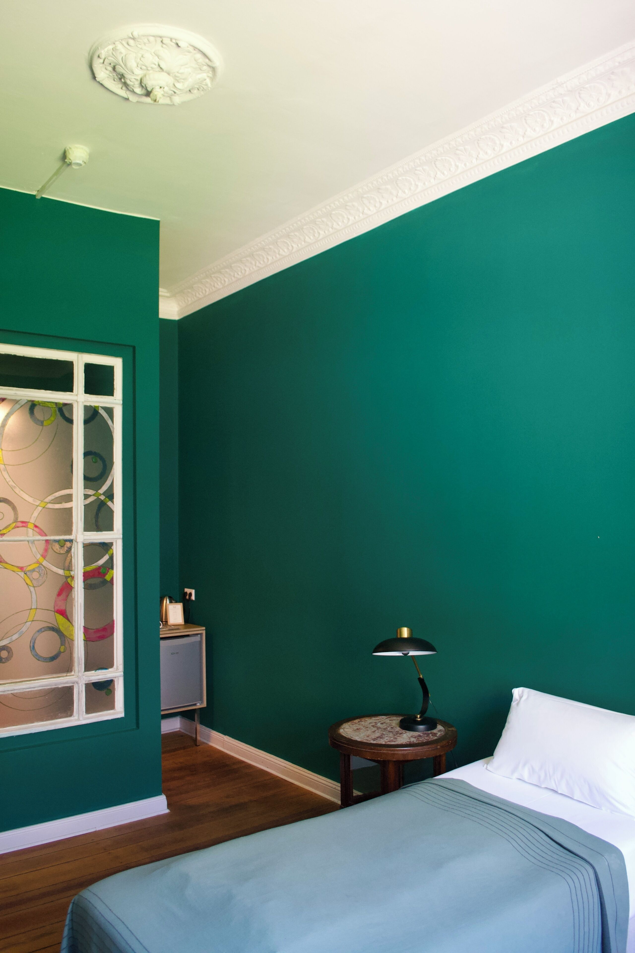 A room with teal walls, a bed with a white pillow, a small round table with a lamp, and a window with decorative glass.