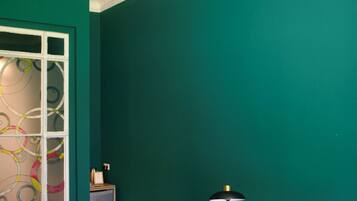 A room with teal walls, a bed with a white pillow, a small round table with a lamp, and a window with decorative glass.