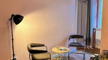 Una habitación con piso de madera, dos sillas, una mesita redonda con una revista, una lámpara de pie y un radiador.