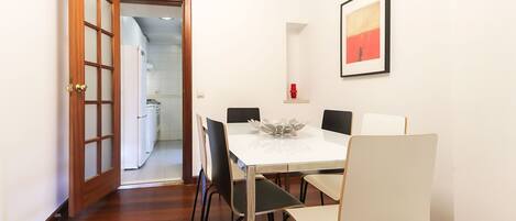 Un comedor con una mesa blanca y sillas negras, un cuadro enmarcado en la pared, y una puerta de madera que da a la cocina.