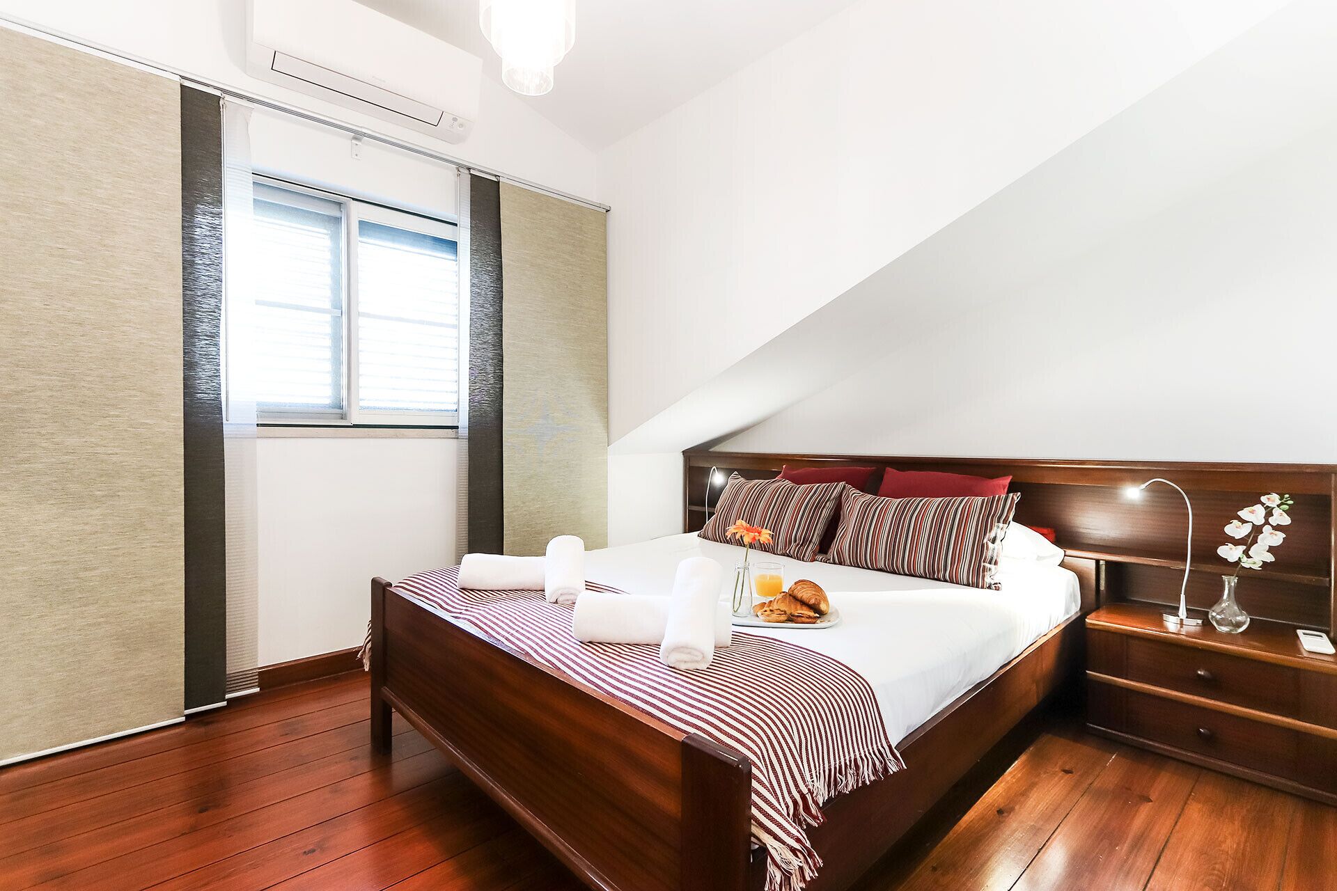 A bedroom with a wooden bed, a bedside table, a nightstand, a lamp, and a window with blinds.