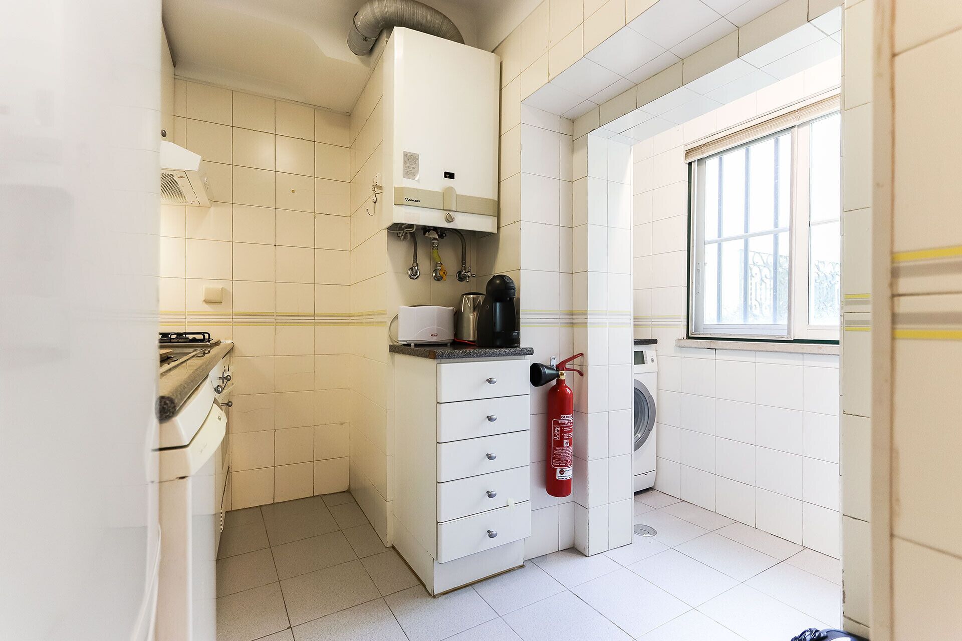 A compact kitchen with a washing machine, a fire extinguisher, and a window providing natural light.