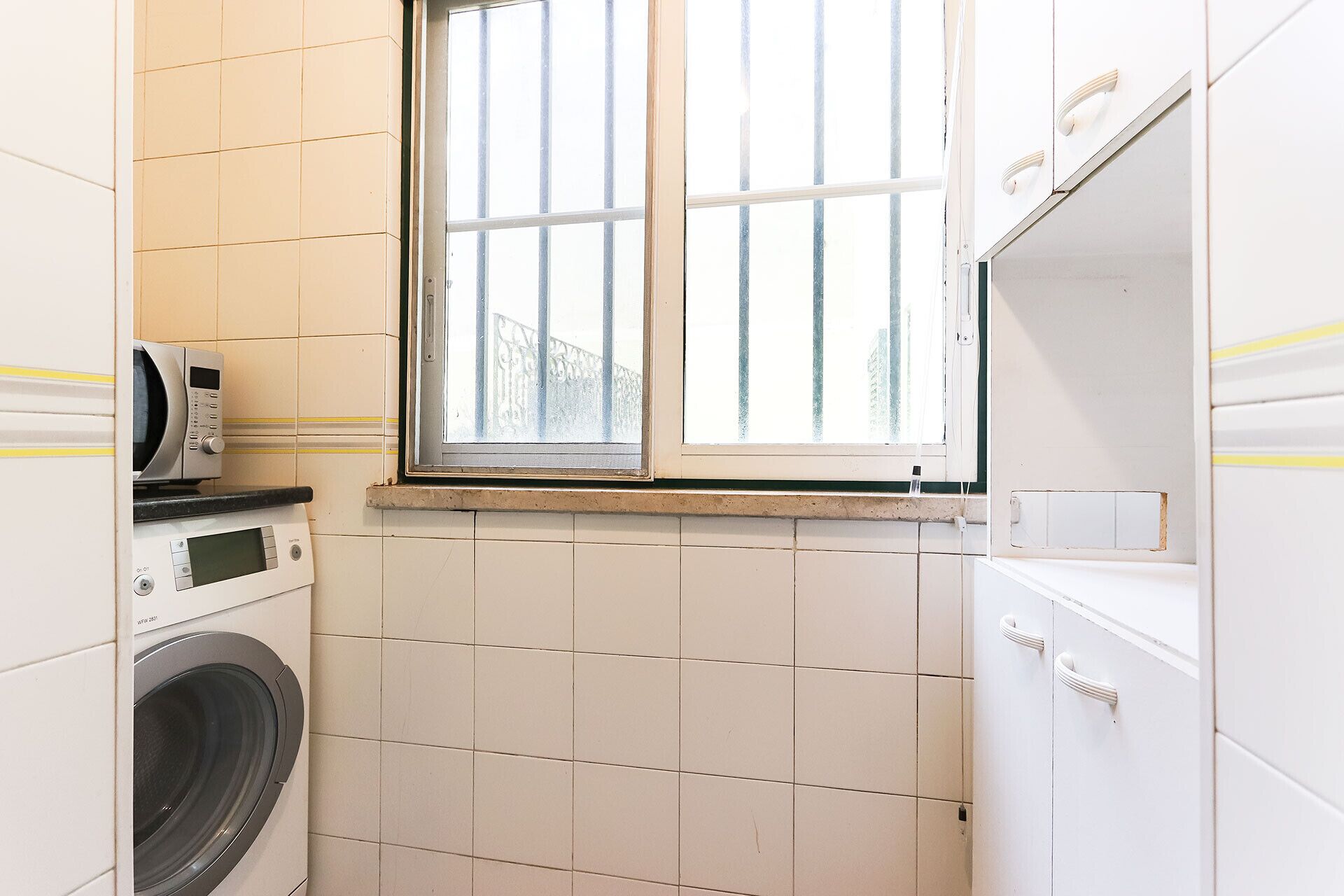 A laundry room with a washing machine, a microwave, and a window with bars.