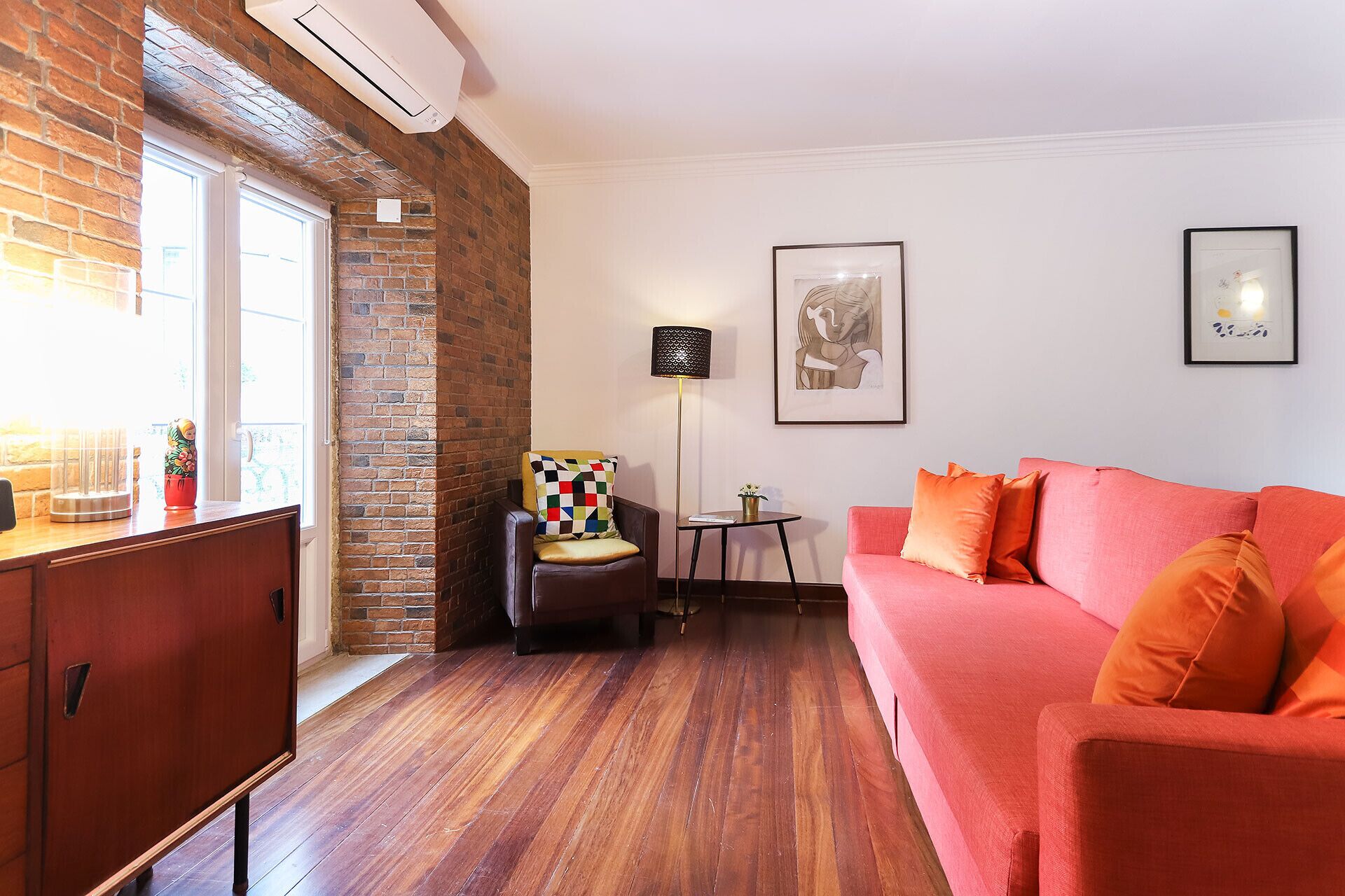 A cozy living room with a brick accent wall, a wooden floor, a red sofa, a black armchair, and a small table with a lamp.