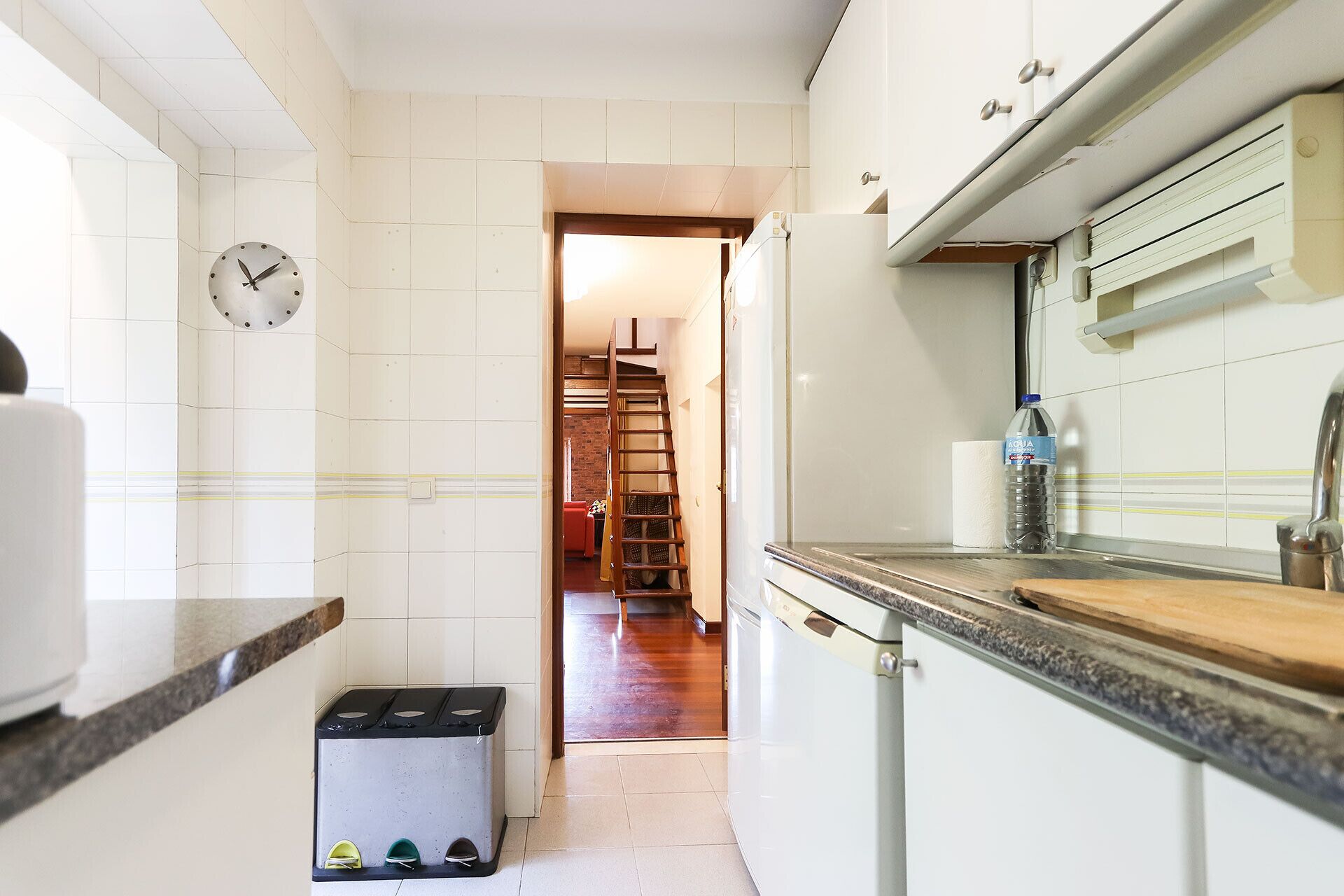 Eine schmale Küche mit weißen Schränken, einer Edelstahl-Spüle und einer Holz Arbeitsplatte. Ein Mülleimer steht auf dem Boden, und eine Wasserflasche liegt auf der Arbeitsplatte.