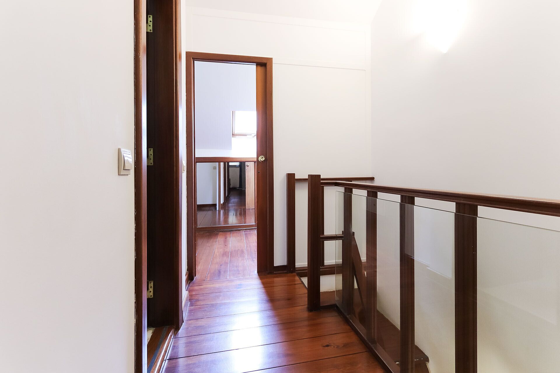 A narrow wooden hallway with a glass railing and a door leading to another room.