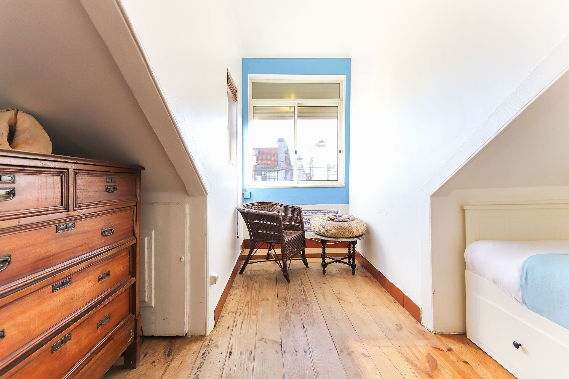 A narrow hallway with a wooden dresser, a wicker chair, a small round table, and a bed with a blue cover.