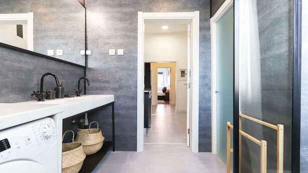 A modern bathroom with a large mirror, a white sink, and a black faucet. There are three woven baskets on the counter, and a washing machine is visible in the corner. - Rato Design With Terrace By Homing (Lisbon)