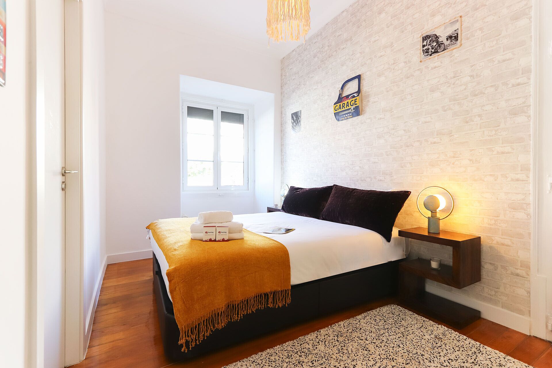 A bedroom with a bed, a window, a wall-mounted light, and a small table with a candle.
