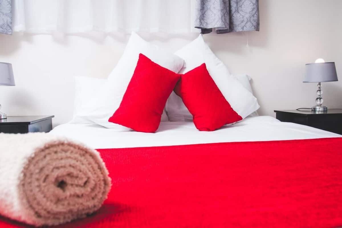 Una cama bien hecha con almohadas blancas y rojas, una colcha roja y una manta beige enrollada en el suelo.