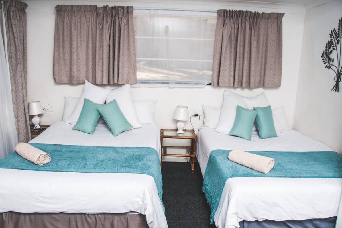 Una habitación de hotel con dos camas, cada una con una colcha y almohadas color turquesa, una mesita de noche pequeña con una lámpara, y una ventana con cortinas.