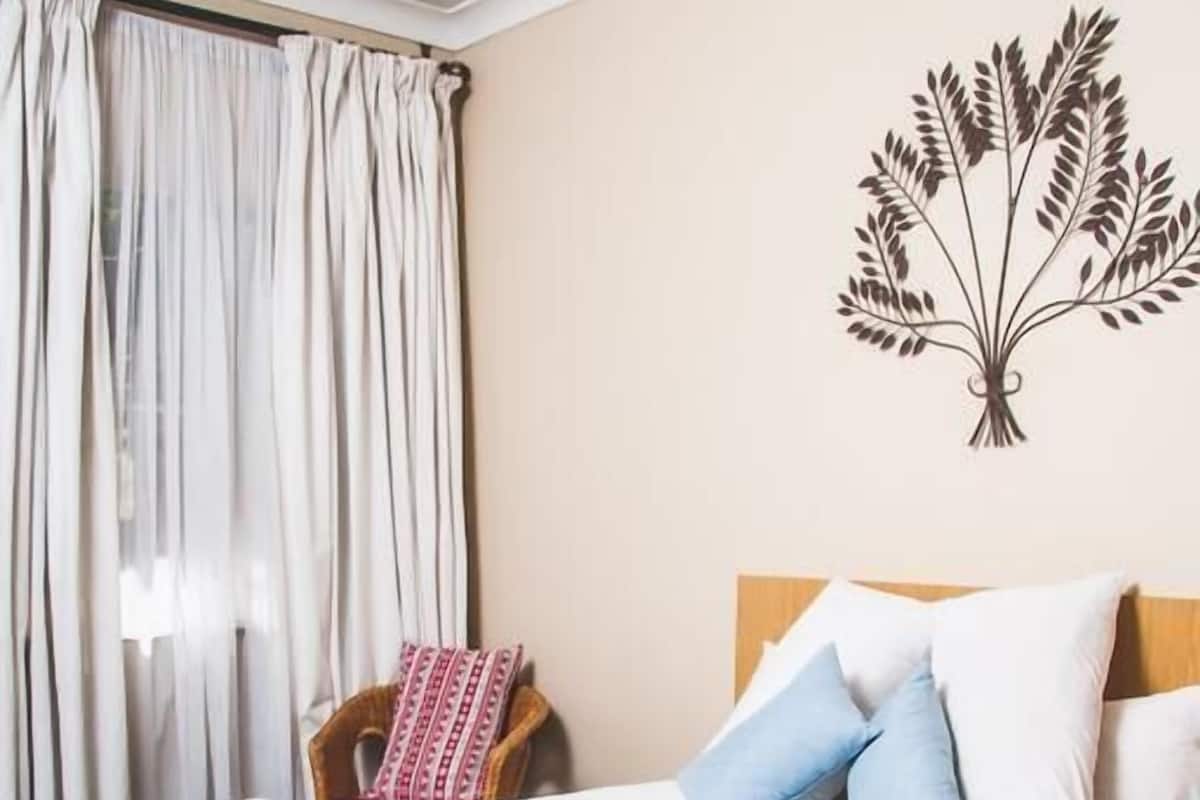 Una cama bien hecha con una colcha azul, almohadas blancas y un cabecero. Hay una silla con un cojín rojo y blanco, y una decoración de pared con un árbol.