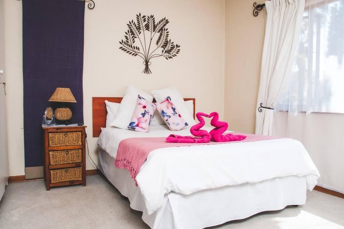Un dormitorio con una cama, almohadas, un adorno en la pared, una mesita de noche, una lámpara y una ventana con cortinas.