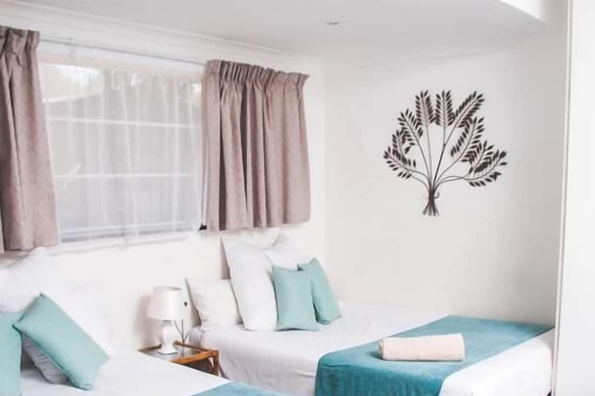 Habitación de hotel con dos camas, una ventana con cortinas, un adhesivo en la pared y una mesita de noche con lámpara.