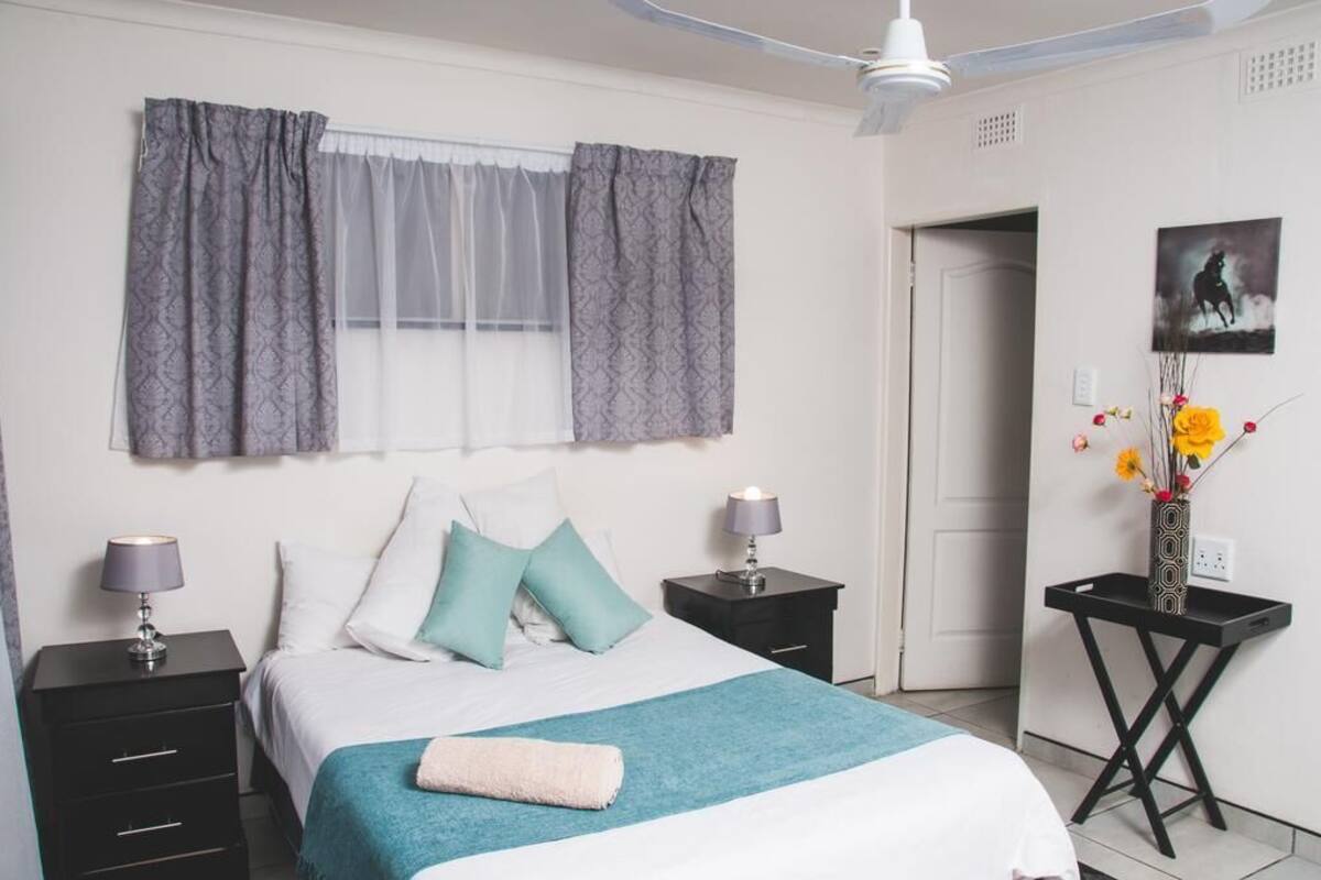 Un dormitorio con una cama, dos mesitas de noche, una mesilla auxiliar, un ventanal con cortinas, una obra de arte colgada en la pared y una pequeña mesa con un florero.