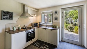 Cocina compacta con armarios blancos, estufa, microondas y ventana con vistas a un jardín.
