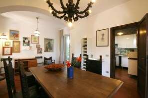 A dining area with a wooden table, chairs, and a chandelier. There are framed pictures on the walls and a bookshelf in the background.