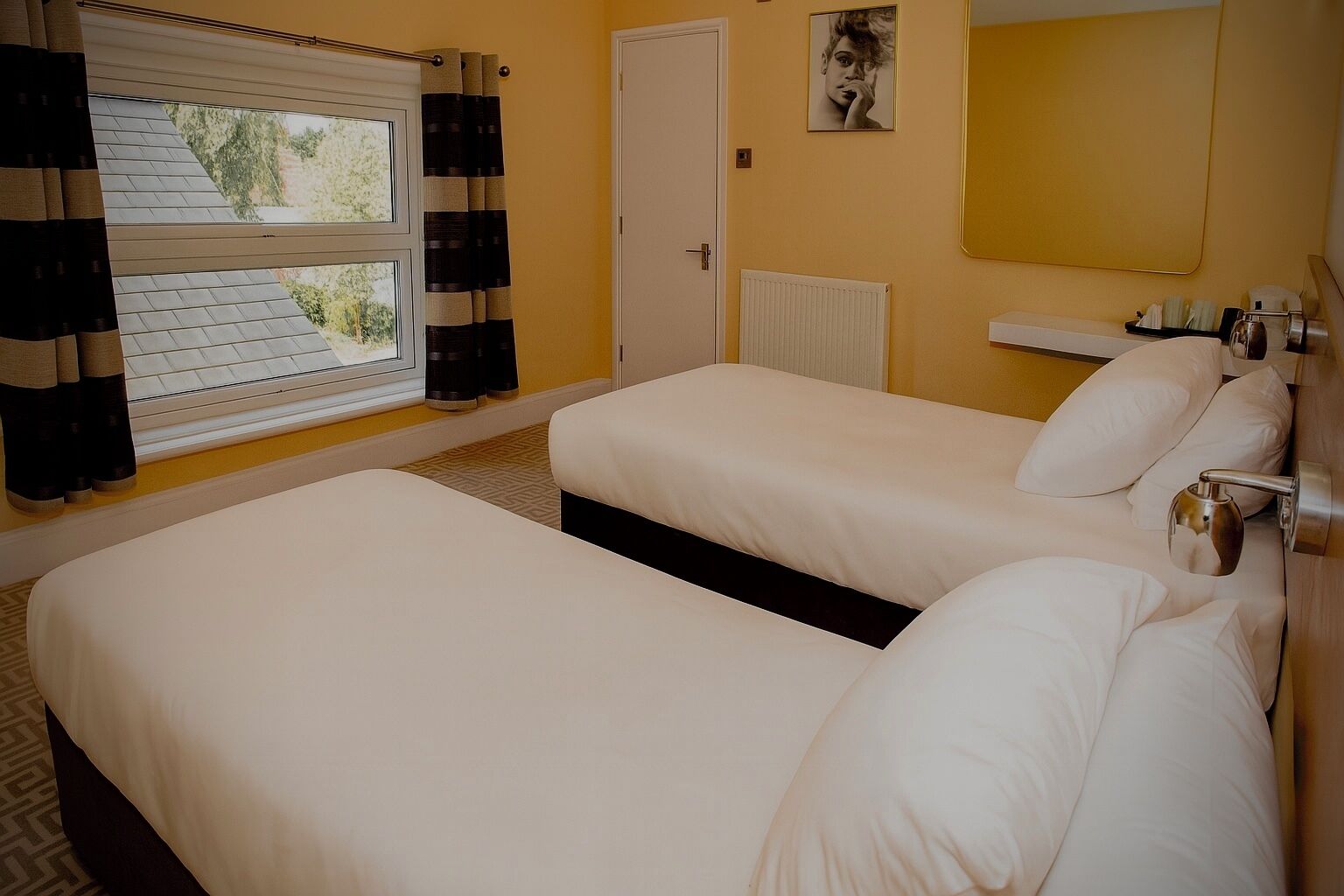 A hotel room with two beds, a window with curtains, a radiator, and a bedside table with a lamp.