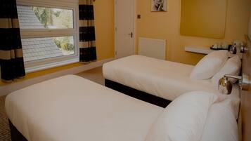 A hotel room with two beds, a window with curtains, a radiator, and a bedside table with a lamp.