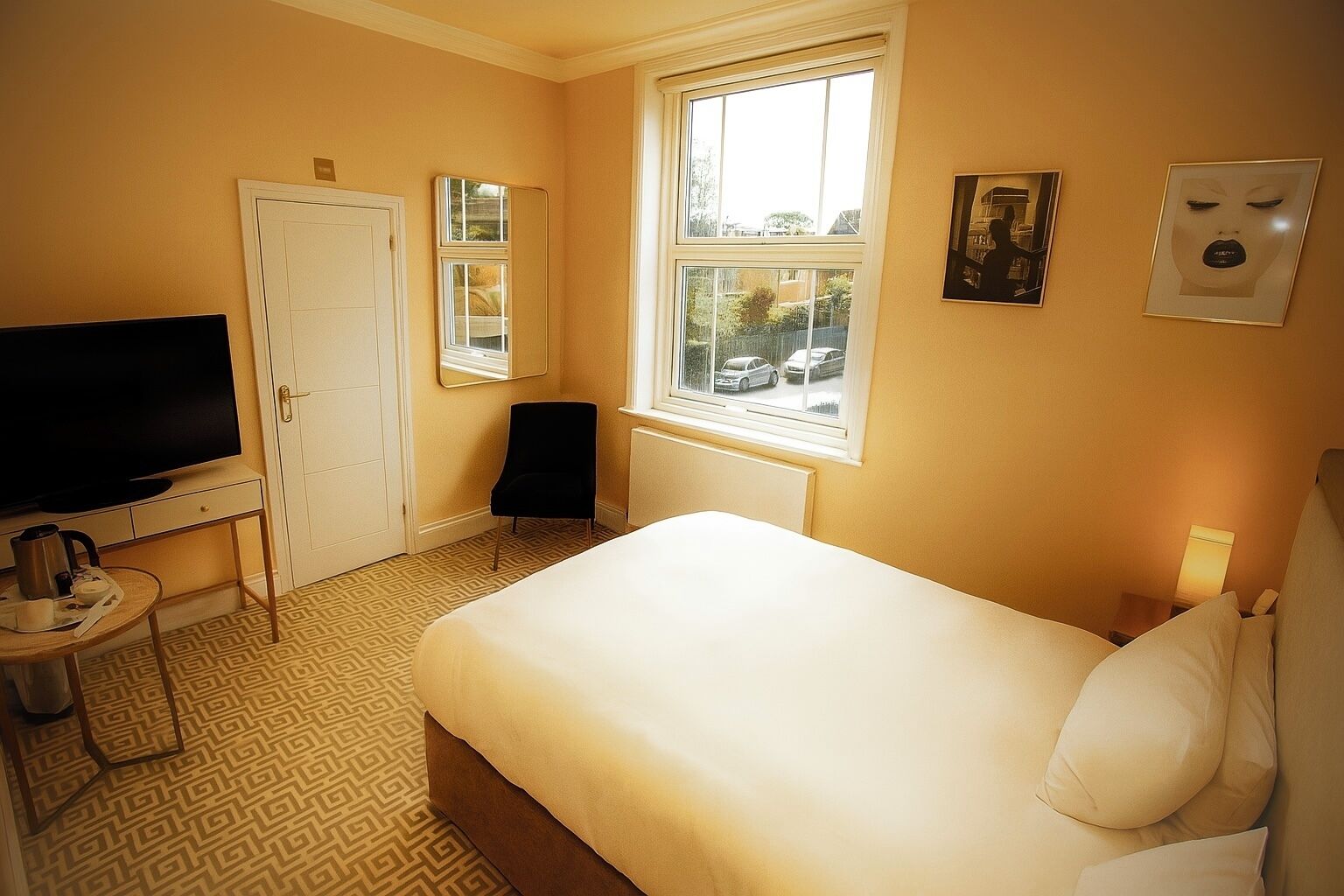 A hotel room with a bed, a TV, a chair, a desk with a kettle, and a window with a view of cars.