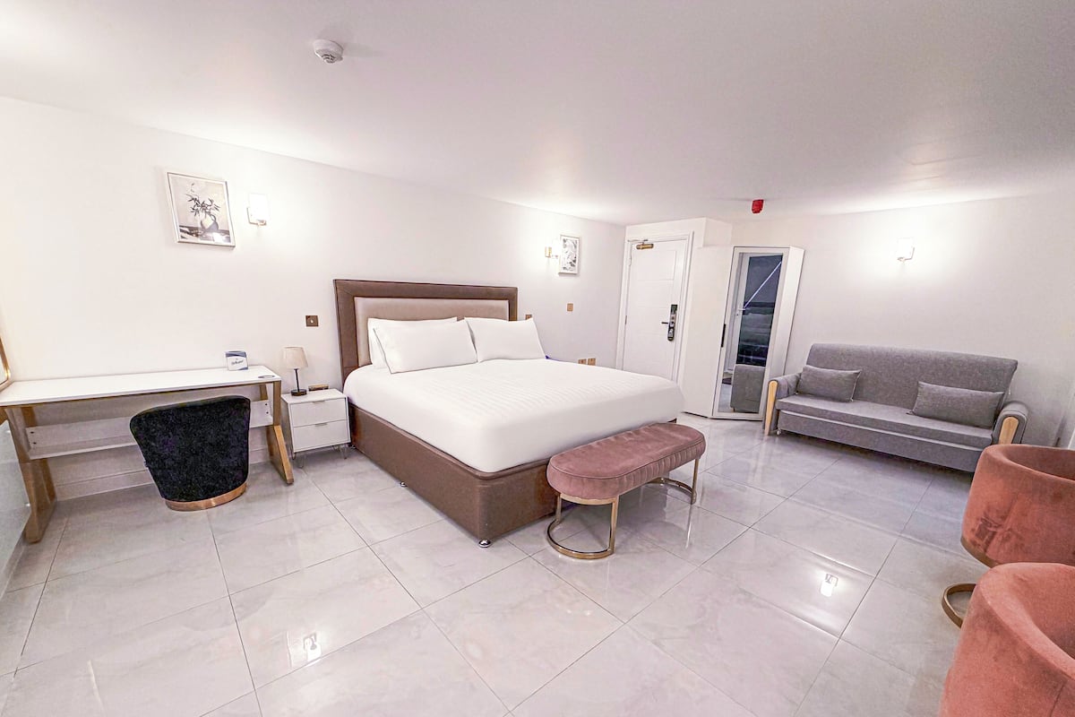 A modern hotel room with a large bed, a desk, a grey sofa, and a pink armchair.