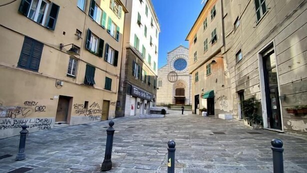 A narrow street with stone pavement and bollards, flanked by buildings with graffiti and a church with a circular window. - Splendida Dimora Negri (Genoa)
